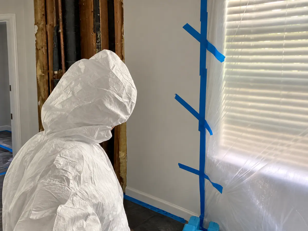 Mold remediation technician in a protective suit working near exposed wall framing and plastic containment sheeting.